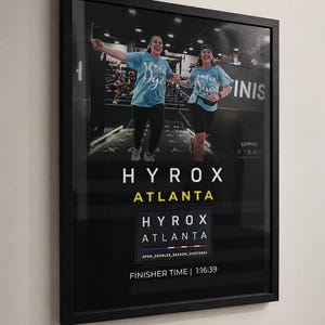 May include: A black framed poster featuring a photograph of two people running in light blue shirts at a Hyrox event. The text reads HYROX ATLANTA, with a smaller box containing HYROX ATLANTA OPEN_DOUBLES_SEASON_2025/2024 and FINISHER TIME | 1:16:39.
