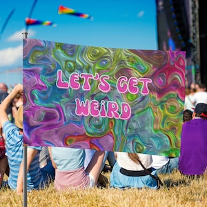 Puede incluir: Una bandera con el texto "LET'S GET WEIRD" en letras rosas sobre un fondo colorido y arremolinado de azules, verdes y morados. La bandera se sostiene en un festival de música, con gente sentada en la hierba en primer plano.