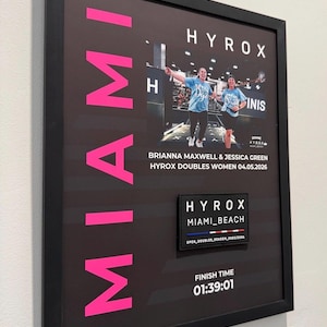 May include: Framed print featuring two women in blue shirts, celebrating a HYROX doubles event. The word "MIAMI" is in large pink letters. Includes event details: Brianna Maxwell & Jessica Green, HYROX Doubles Women 04.05.2026, and a finish time of 01:39:01.