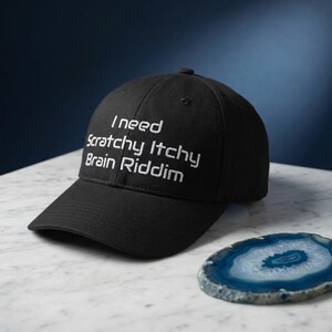 May include: Black baseball cap with the embroidered text "I need Scratchy Itchy Brain Riddim" in white. The cap is on a white marble surface with a blue and white agate coaster.