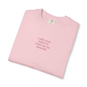 May include: A pale pink t-shirt with the phrase "I sold some tickets to come see my downfall" printed in a darker pink. The shirt is folded neatly, with a label visible on the collar.