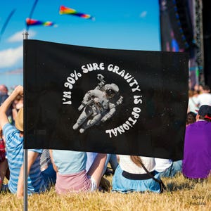 Pode incluir: Bandeira preta com o texto "I'm 90% sure gravity is optional" em um design circular em torno de uma ilustração de astronauta. A bandeira está em um mastro, com pessoas e pipas ao fundo.
