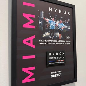 May include: Framed print with the word "MIAMI" in pink. It features a photo of two women in blue shirts, the text "HYROX DOUBLES WOMEN 04.05.2026", and a "HYROX MIAMI_BEACH" patch. The finish time is 01:39:01.