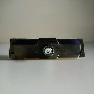 Puede incluir: Un objeto metálico gris oscuro con una lente circular en el centro. El objeto rectangular tiene una sección central ligeramente elevada y dos pequeños agujeros en cada extremo. La lente tiene un centro blanco y un anillo plateado.