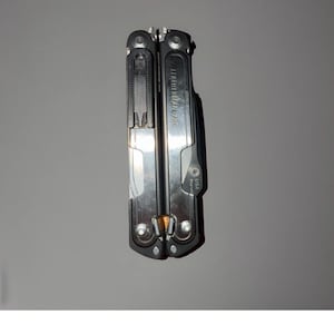 以下が含まれることがあります： 「LEATHERMAN ARC」の文字が見える、シルバーと黒のマルチツール。ナイフの刃、ペンチ、その他の道具が付いています。ツールは閉じられており、金属製のように見えます。