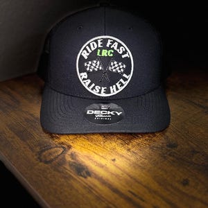 May include: Black trucker hat with a white and green embroidered design. The design features the words "RIDE FAST RAISE HELL" in a circular pattern, with two crossed checkered flags in the center. The hat has a curved brim and a mesh back.