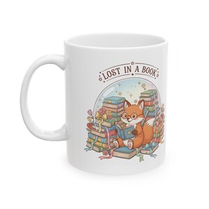 Könnte beinhalten: Weißer Keramikbecher mit einem Cartoon-Fuchs, der ein Buch liest, eine Brille trägt und von Büchern umgeben ist. Der Becher trägt den Text "LOST IN A BOOK" in einem kreisförmigen Design. Das Design umfasst Blumen, Bänder und Sterne.