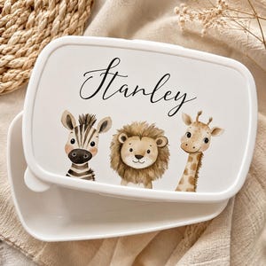 May include: A white rectangular lunch box with a lid featuring the name Stanley in black script. The lid displays watercolour illustrations of a zebra, a lion, and a giraffe arranged in a row. The container is open, showing a matching white interior.