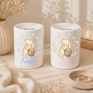 May include: Two white ceramic cylindrical money boxes featuring a Winnie the Pooh illustration with a honey pot inside a floral wreath. One box has the name Frankie in blue script, and the other has the name Eliza in pink script.