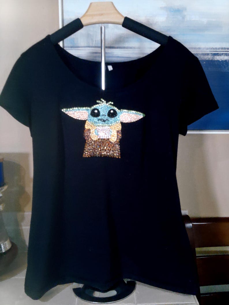 May include: Black short-sleeve t-shirt featuring a rhinestone design of a cartoon character. The design includes light blue, tan, and brown colors. The shirt is hanging on a wooden hanger.