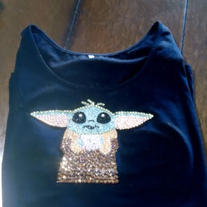May include: Black t-shirt featuring a rhinestone design of a character with large ears and a tan and gold sequined outfit. The character's face is light blue with black eyes and a small nose. The t-shirt is folded on a wooden surface.