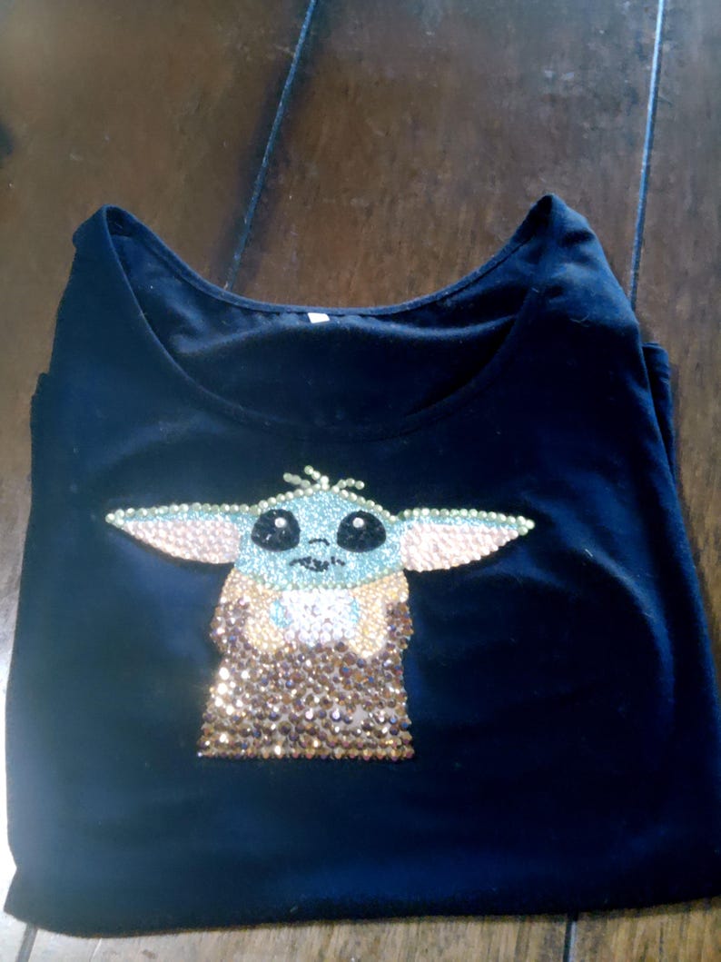 May include: Black t-shirt featuring a rhinestone design of a character with large ears and a tan and gold sequined outfit. The character's face is light blue with black eyes and a small nose. The t-shirt is folded on a wooden surface.