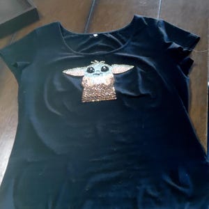 May include: Black t-shirt with a scoop neck featuring a rhinestone design of a character with large ears and large eyes. The character's face is light blue and brown, with a brown and gold body.