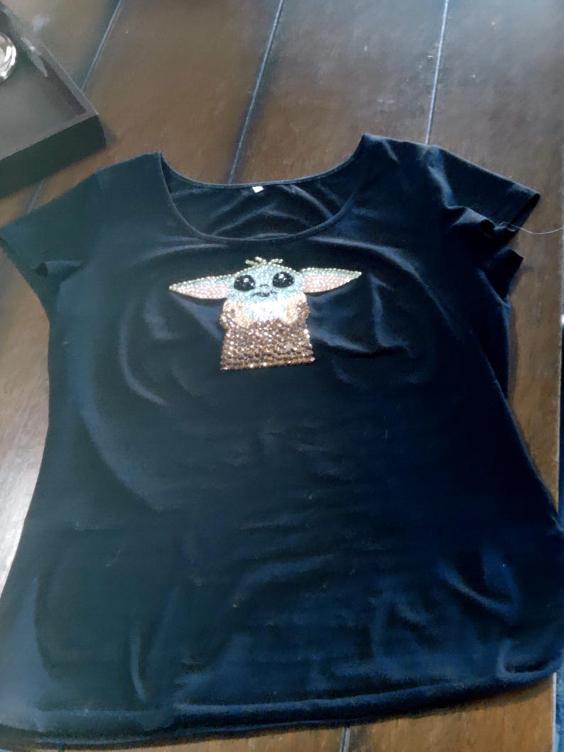 May include: Black t-shirt with a scoop neck featuring a rhinestone design of a character with large ears and large eyes. The character's face is light blue and brown, with a brown and gold body.