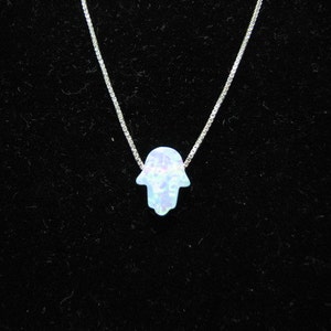 May include: A delicate silver necklace featuring a pendant in the shape of a Hamsa hand. The pendant has a light blue opal-like appearance. The necklace is displayed against a black background.