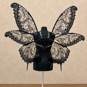 May include: A pair of large, black butterfly wings featuring intricate, lace-like cutouts and swirling patterns. The wings are attached to a central black harness frame with silver-toned metal hardware, including clips and adjustable straps.