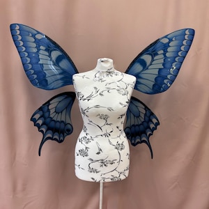 Op de afbeelding: Een witte paspop met een bloemenpatroon, versierd met grote blauwe vlindervleugels. De vleugels hebben een gedetailleerd ontwerp met verschillende blauwtinten. De paspop is gemonteerd op een zilveren standaard, tegen een roze achtergrond.