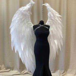 Puede incluir: Un vestido negro sin tirantes con silueta de sirena se exhibe en un maniquí, combinado con grandes alas de ángel blancas de plumas. Las alas están colocadas detrás del vestido, creando un efecto dramático. El vestido tiene una abertura frontal.