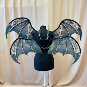 Könnte beinhalten: Fledermausflügel mit schwarzem Geschirr, ausgestellt auf einer schwarzen Schaufensterpuppe. Die Flügel sind ein Farbverlauf aus Blau und Grau, mit schwarzem Spitzensaum und weißen floralen Spitzendetails. Die Flügel sind zum Tragen konzipiert.