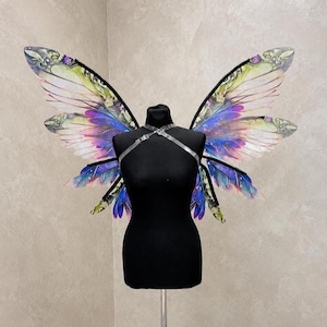 Può includere: Un manichino nero con ali di farfalla iridescenti. Le ali presentano un design colorato con sfumature di viola, blu, verde e giallo. Le ali sono attaccate al manichino con un'imbracatura trasparente. Il manichino è su un supporto.