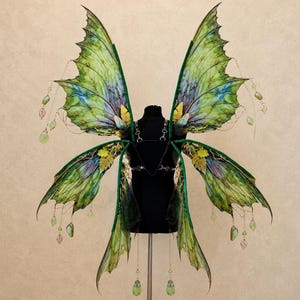 May include: Large decorative wings featuring a vibrant green and blue gradient pattern with intricate vein detailing. The wings are adorned with dangling light green teardrop beads and small gold leaf charms, attached to a black harness frame.