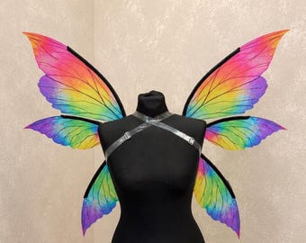May include: Rainbow-colored butterfly wings with black outlines and a clear harness are displayed on a black mannequin. The wings feature vibrant hues of pink, orange, yellow, green, and purple, creating a colorful and whimsical effect. The wings are designed for costume or cosplay.