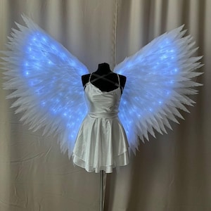 May include: A white satin dress with a layered skirt is displayed on a mannequin, paired with large, feathery white angel wings. The wings are illuminated with blue LED lights, creating a glowing effect. The outfit is designed for costume or cosplay.