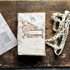 May include: A handmade journal featuring a collage cover with floral patterns, an acorn illustration, and the text autumn days. The spine is decorated with cream-colored lace trim, and a long, cream-colored fabric ribbon with black polka dots is attached.