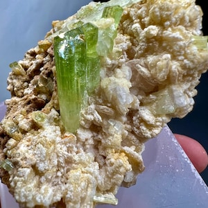 Pode incluir: Uma amostra mineral em close-up com cristais verdes vibrantes emergindo de uma matriz bege. Os cristais são translúcidos e vítreos. A matriz tem uma superfície áspera e texturizada. A amostra é exibida em uma superfície roxa clara.