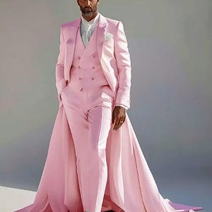 Puede incluir: Un traje de tres piezas rosa claro con una larga cola. El traje incluye un chaleco cruzado, una americana con un prendedor de flores y pantalones a juego. El conjunto se completa con una camisa blanca y botas blancas.