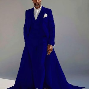 Puede incluir: Un llamativo traje azul cobalto con una cola extendida. El traje presenta una chaqueta a medida con un pa&ntilde;uelo blanco y un pantal&oacute;n a juego. El modelo lleva una camisa blanca y botas blancas. El traje es una audaz declaraci&oacute;n de moda.