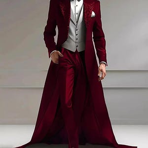 May include: A formal burgundy suit with a long coat, embroidered lapels, a grey waistcoat, and matching trousers. The suit includes a black bow tie and a white pocket square. The coat extends to the ankles, creating a dramatic silhouette.