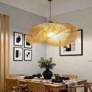 May include: A woven rattan pendant light fixture hangs above a wooden dining table. The light fixture has a unique, organic shape and casts a warm glow. The dining table is set with black plates and a centerpiece of greenery in a black vase.