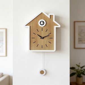 May include: A wooden cuckoo clock in the shape of a house, with a white border. The clock face has black hands and markers. A small bird is visible in the center. A white pendulum hangs below. The clock is mounted on a white wall.