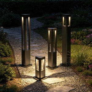 Può includere: Quattro luci per sentieri esterni nere con un design quadrato sono illuminate in un giardino. Le luci sono di altezze diverse, con un bagliore caldo che emana dall'interno. Le luci sono posizionate lungo un sentiero di pietra.