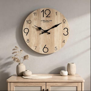 Puede incluir: Un reloj de pared redondo de madera con números y manecillas negras. La esfera del reloj tiene un patrón de veta de madera clara. El texto "est 1765 vintage cloche QUARTZ CLOCK" está impreso en la esfera. El reloj está sobre un mueble de madera.
