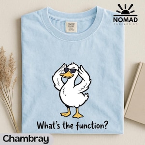 Puede incluir: Camiseta azul claro con un pato de dibujos animados con gafas de sol y el texto "What's the function?". La camiseta tiene cuello redondo y una etiqueta "Comfort Colors". El logotipo de la marca "Nomad Threads St" es visible.