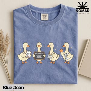 May include: Blue jean colored t-shirt with a crew neck. The shirt features a graphic of four cartoon ducks, each holding different items. The text "ABCs of Behavior: Antecedent Behavior Consequence" is on a chalkboard. The brand logo is in the upper right.
