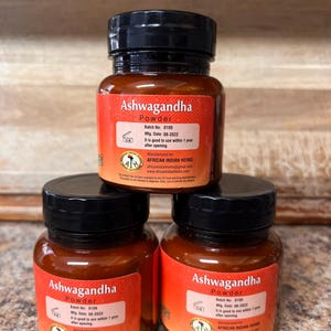 May include: Three small glass jars of Ashwagandha powder are stacked. Each jar has a black lid and a red label with white text. The labels include the text "Ashwagandha Powder", batch and manufacturing information, and the manufacturer's name.