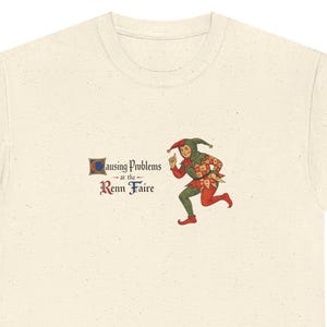 May include: Cream-colored t-shirt featuring a whimsical illustration of a jester carrying a bundle of items. The text "causing problems at the Renn Faire" is printed on the shirt. The jester is depicted in green, red, and gold.
