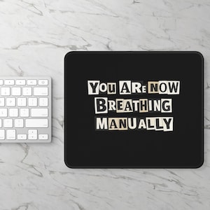 May include: A black rectangular mousepad with the text "You Are Now Breathing Manually" in a cut-out letter style. A white keyboard is partially visible on the left side of the image, set against a marble-patterned surface.