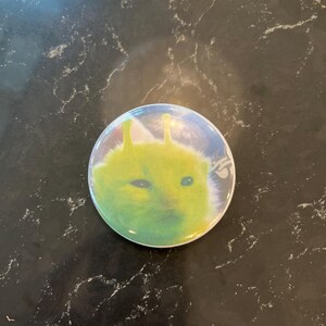 May include: A round button featuring a green alien face with large eyes and two antennae. The alien has a slightly sad expression. The button has a glossy finish and is set against a dark background.
