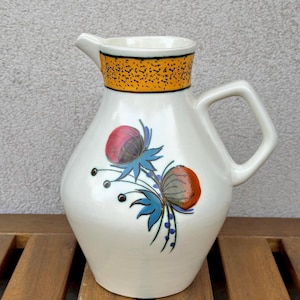 Może przedstawiać: Biały ceramiczny dzban z dzióbkiem i uchwytem. Dzbanek posiada dekoracyjny kwiatowy wzór w kolorze czerwonym, niebieskim i pomarańczowym. Obrzeże jest zaakcentowane żółto-czarnym wzorem. Dzbanek stoi na drewnianej powierzchni.