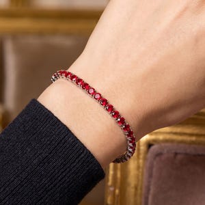 Peut inclure: Un bracelet tennis composé d'une rangée continue de pierres précieuses rondes d'un rouge éclatant, serties sur une bande en métal argenté poli. Le bracelet est porté au poignet, mettant en valeur la taille uniforme et la coupe ronde de chaque pierre.