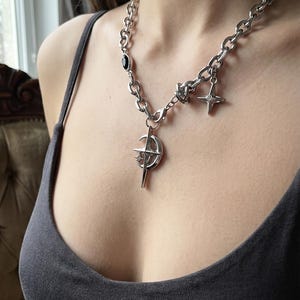 Peut inclure: Un collier à chaîne argentée doté d'un pendentif central en forme de croissant de lune et d'étoile à quatre branches. La chaîne comprend une petite perle ovale noire et une breloque étoile à quatre branches. Les maillons sont épais et arrondis.