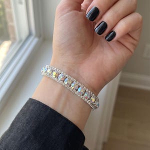 Peut inclure: Un bracelet flexible composé d'une rangée unique de cristaux facettés iridescents et multicolores sertis dans une monture en métal argenté. Les cristaux présentent un spectre de couleurs incluant le rose, le bleu et le jaune, reflétant la lumière.