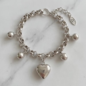 Op de afbeelding: Een zilveren schakelarmband met een centrale hartvormige bedel en vier kleinere bolvormige bedels. De armband is voorzien van een karabijnsluiting en een klein ovaal label met de tekst 925, wat duidt op de constructie van sterling zilver.