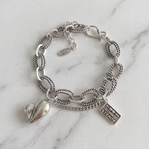Op de afbeelding: Een zilveren kettingarmband met een mix van gedraaide en gladde ovale schakels. De armband bevat een gepolijste hartvormige bedel, een rechthoekig plaatje met de tekst GOING STEADY en een klein ovaal plaatje met de tekst Silver 925.