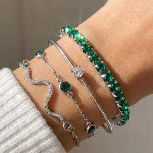 Peut inclure: Un ensemble de quatre bracelets argentés. L'un présente une rangée de pierres rondes vert émeraude, un autre est un jonc lisse avec une pierre claire, le troisième est une chaîne fine avec des accents verts et le dernier est un bracelet ondulé.