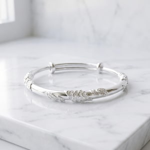 Op de afbeelding: Een zilverkleurige armband met een verstelbaar schuifmechanisme en een centraal bloemmotief met reliëfblad-details. Het metalen oppervlak is gepolijst en reflecterend, met een delicaat botanisch ontwerp langs de ronde band.